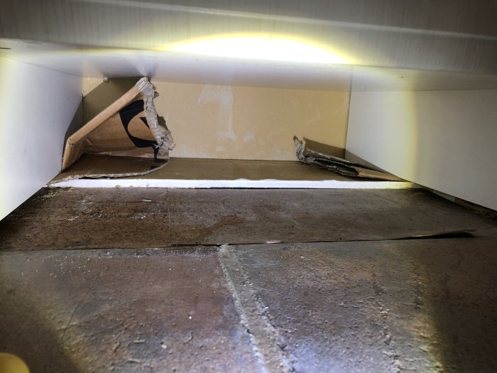 Glipor och hål under köksskåp med synlig tegelvägg och rostiga skruvar, tecken på drag och fuktskador.