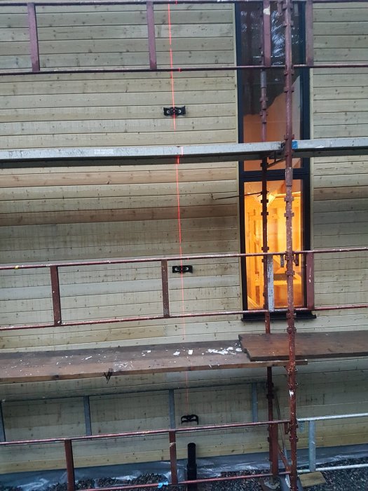 Byggnadsställning framför en träbeklädd fasad med en korslaser som projicerar röda linjer, och hörnfönster upplyst inifrån.