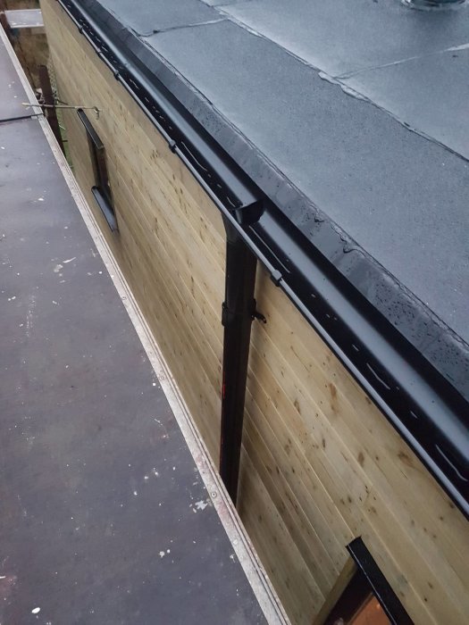 Svarta Lindab-rännor monterade på en byggnad med träpanel, med ett stuprör placerat centralt på väggen.