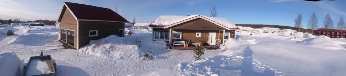 Panoramavy över hus och trädgård omgiven av höga snöhögar under en solig dag.