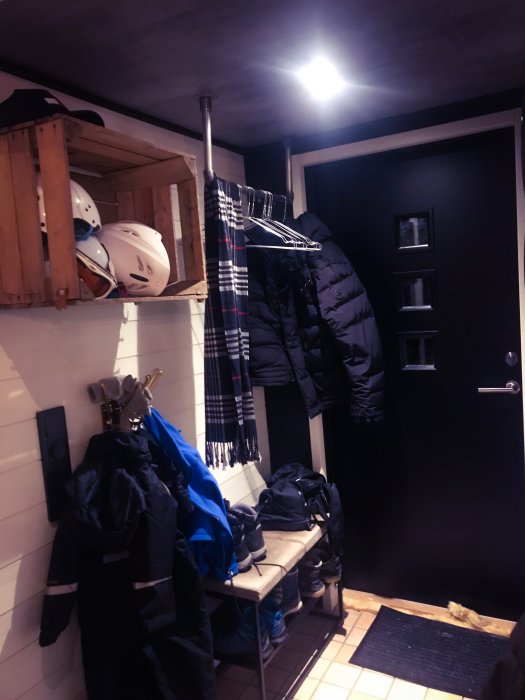 Nyinredd hall med rörhängare för kläder, hylla med skidhjälmar och en sittbänk med skor undertill.