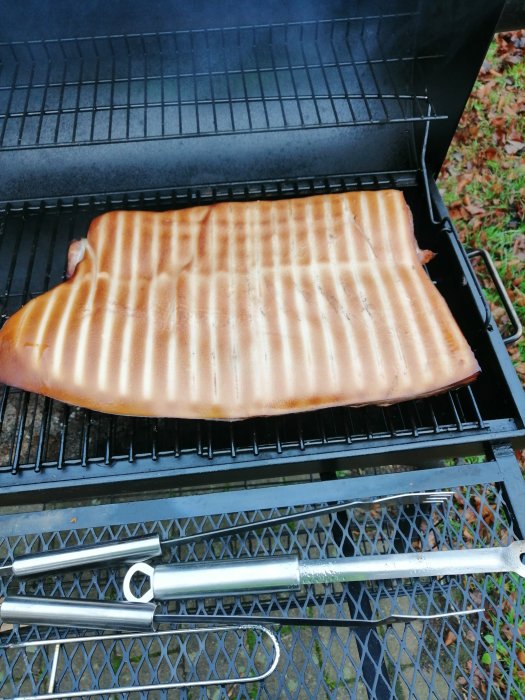 En hel sida av fläskkött med grillränder på en öppen kolgrill med grillverktyg under.