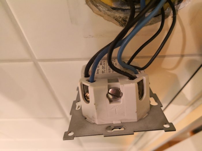 Öppen eluttagsdosa med anslutna kablar under installation på en vit kaklad vägg.