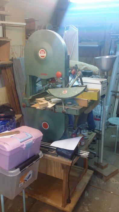 En äldre modell av bandsåg märkt med 'EJCA' i ett rörigt snickeri med verktyg och byggmaterial runtomkring.
