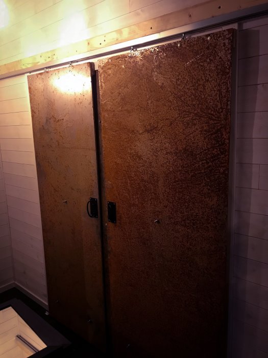 Två rostiga skjutdörrar i metall med svarta handtag, monterade på en vit vägg och upplysta ovanifrån.