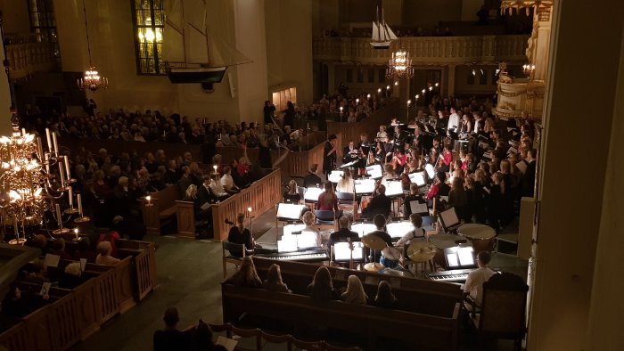 Orkester och kör framför musik inför publik i en upplyst kyrksal under kvällstid.