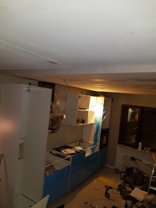 Renoveringsarbete i kök med blå köksluckor och lådor på plats, elinstallationer ej klara, rörigt.