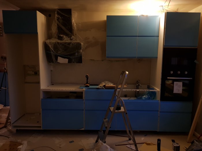 Renovering pågår i kök med blå skåpsluckor, installerade vitvaror och en stege framför diskbänken.