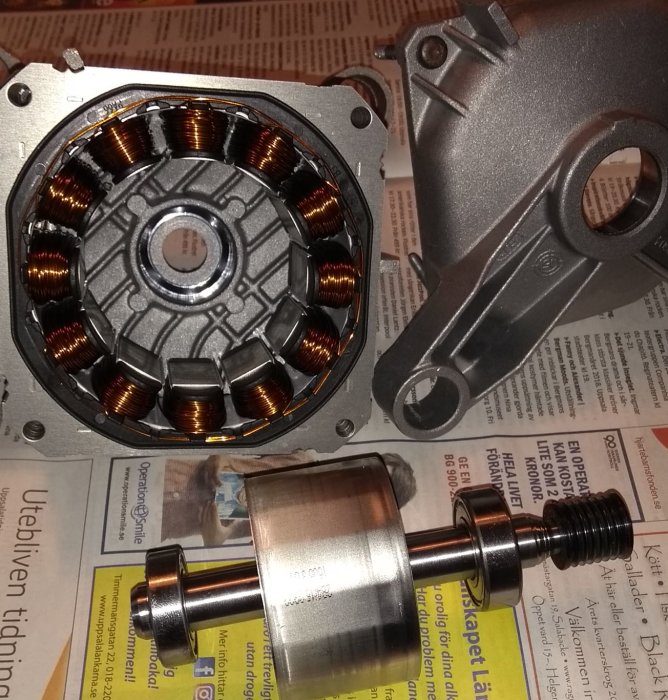 Delar av en elektrisk motor upplagda på en tidning, inklusive stator med kopparlindningar och rotoraxel.