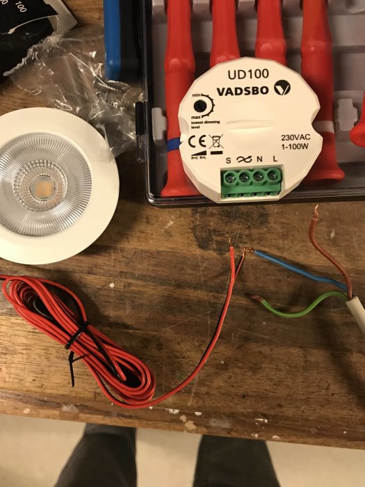 Elektriska kablar och en LED-downlight med en dimmermekanism ovanpå en verktygslåda, placerade på ett träbord.