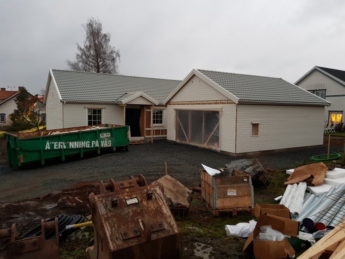 Nybyggt hus med grusat infart och en container, byggmaterial och lerig mark runtomkring.