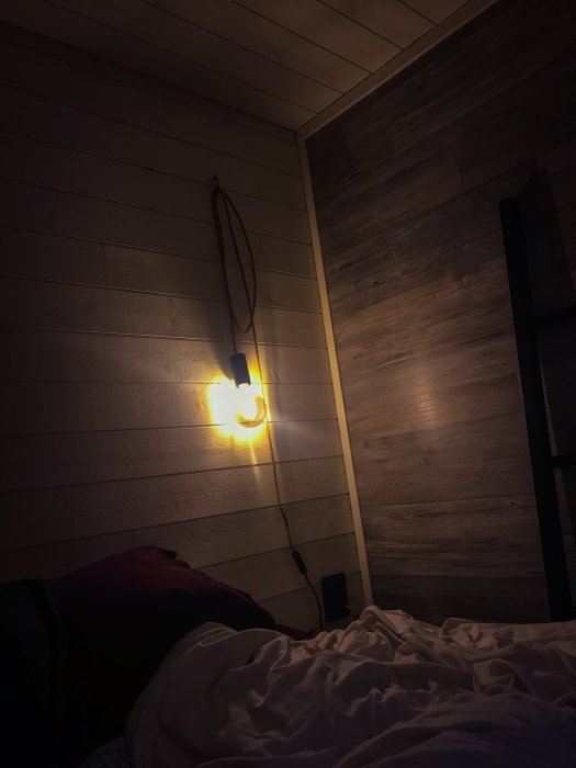 Läslampa monterad på trävägg med mobilhållare och kabel, intill en obäddad säng i halvmörkt sovrum.