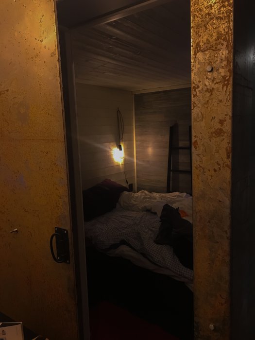 En obäddad säng i ett mysigt sovrum med läslampa på väggen och hängare på en träbalk.