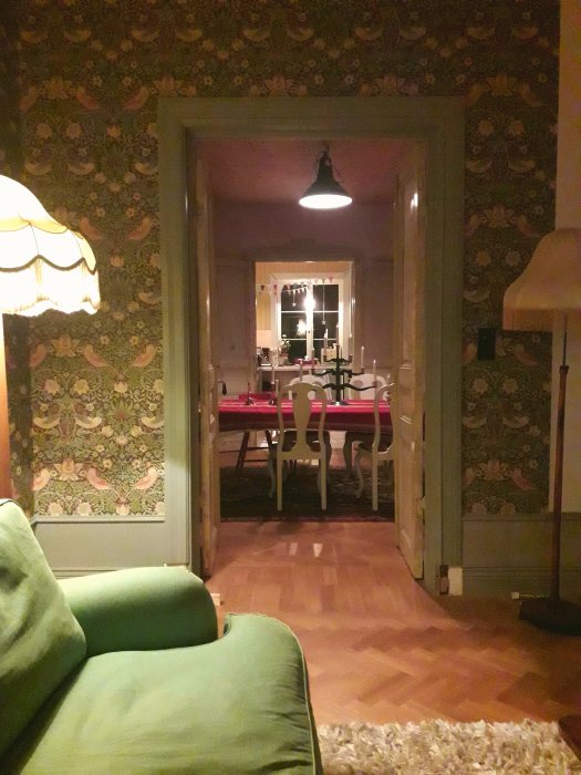 Blick genom en öppen dörr från ett vardagsrum till ett matsal med rött bord och vita stolar, omgiven av vintage tapeter.