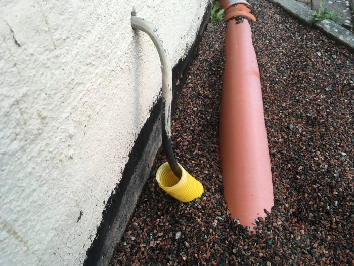 Del av en byggnad med ett synligt avloppsrör och grus nära väggen, med fokus på en tätning vid rörets genomföring.