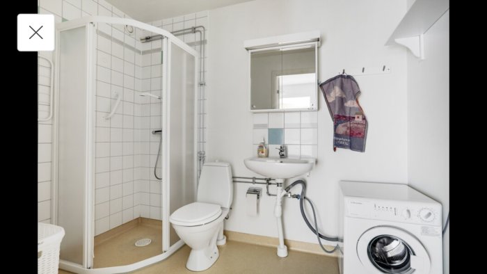 Vitt badrum med duschkabin, toalett, handfat, spegel, tvättmaskin och befintligt stengolv.
