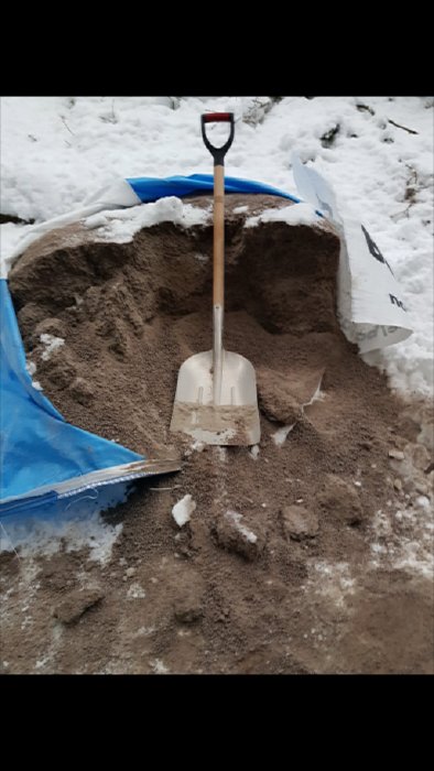 Spade står i en hög med grus bredvid en blå presenning och snötäckt mark, ansträngning vid bygge av hundgård.