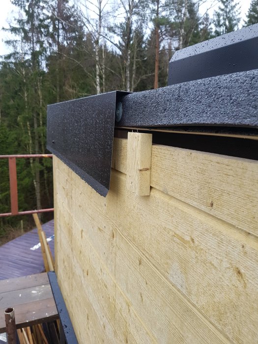 Kröningsplåt monterad på takkant med distansklossar för luftning mot en trävägg, med synliga skruvar och bockade hörn.