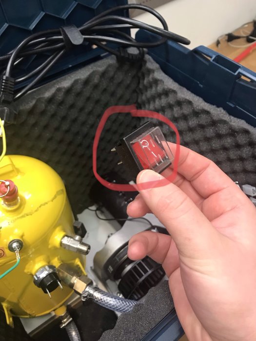 Hand håller en brytare framför öppen l-boxx med en demonterad gul bärbar kompressor och oanslutna elektriska komponenter.