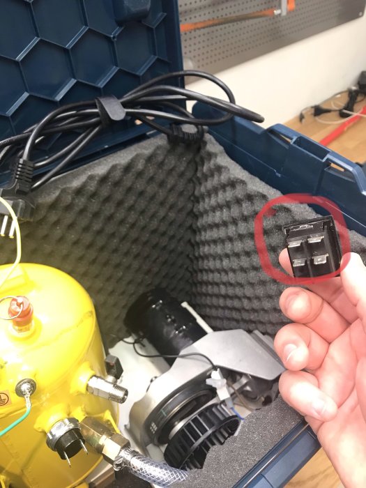 Hand håller en elektrisk kontakt framför en bärbar kompressor och olika komponenter i en blå verktygslåda.