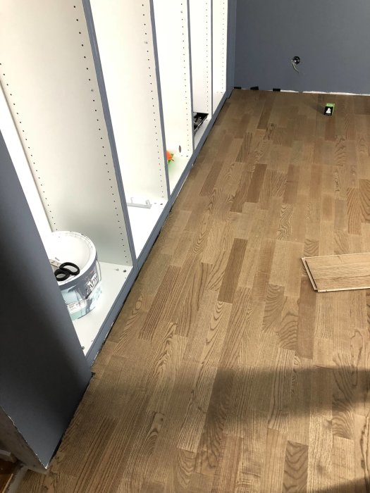Nyinstallerat asklaminatgolv i rum med vita bokhyllor och en färgburk i hörnet.