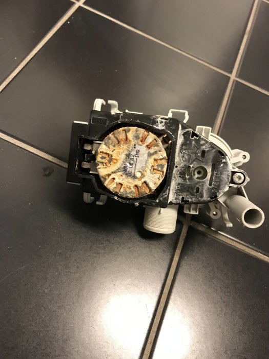 Kalkbelagd och sliten del av en vitvaruprodukt ligger på ett mörkt kakelgolv.