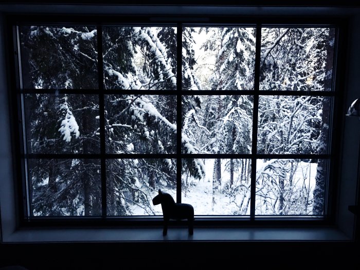 Utsikt från fönster över snötäckta träd i Lindvallen på en vintermorgon med en liten hästfigur på fönsterbrädan.