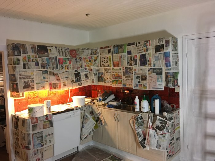 Kök under renovering med väggar och skåpsluckor täckta av tidningspapper, målarburkar och verktyg synliga.