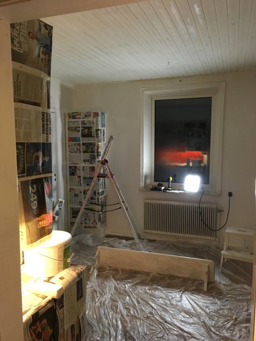 Renovering av kök med tidningar på väggar, plast över golv och en stege, under målningsarbete.