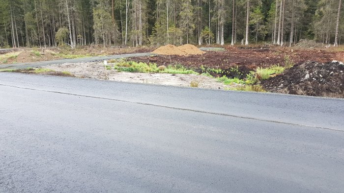 Område med uppgrävda schaktmassor på skogsmark bredvid asfalterad väg, högar av sand och jord.