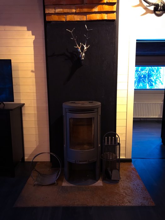 Nyinstallerad kamin med tillbehör framför svart vägg med hjorthuvud och synlig tegel, i nyligen färdigställd skidstuga.