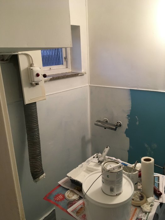 Badrum under renovering med halvmålade väggar, målarburkar och verktyg på golvet.