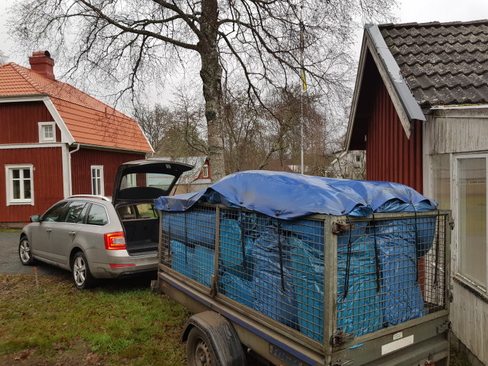 En släpvagn full av blå säckar täckt med presenning bredvid en silverfärgad bil.