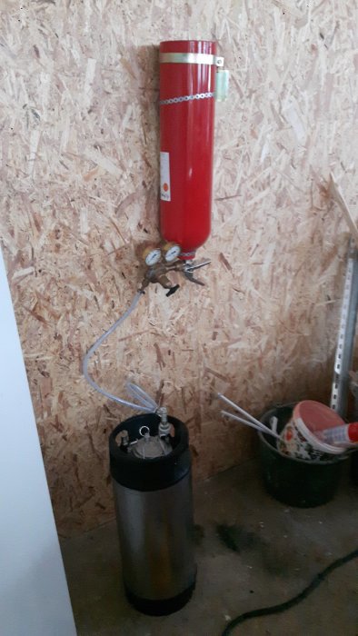 Utrustning för kolsyrning av julöl: röd gasbehållare och svart ölfat mot en OSB-skivvägg.