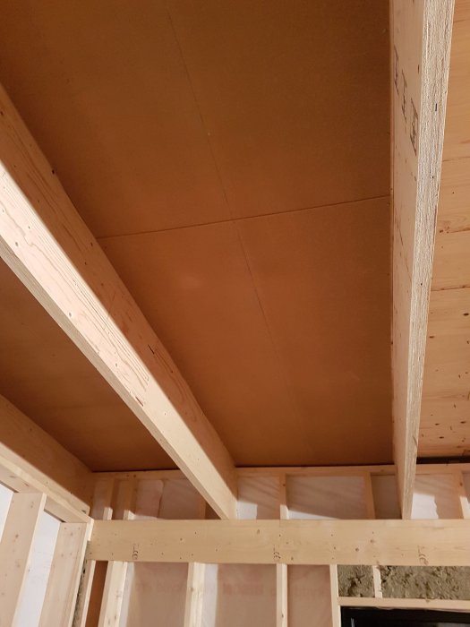 Inre takstruktur i byggprojekt med synliga träbjälkar och nyinstallerad masonitskiva ovanför isolering.