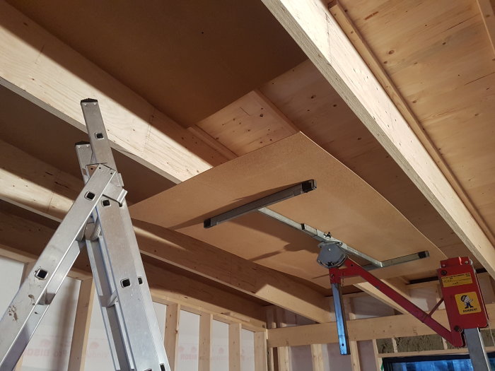 Tak i konstruktion med synliga träbjälkar och en monterad masonitskiva, stegar och en lyftanordning i en byggmiljö.