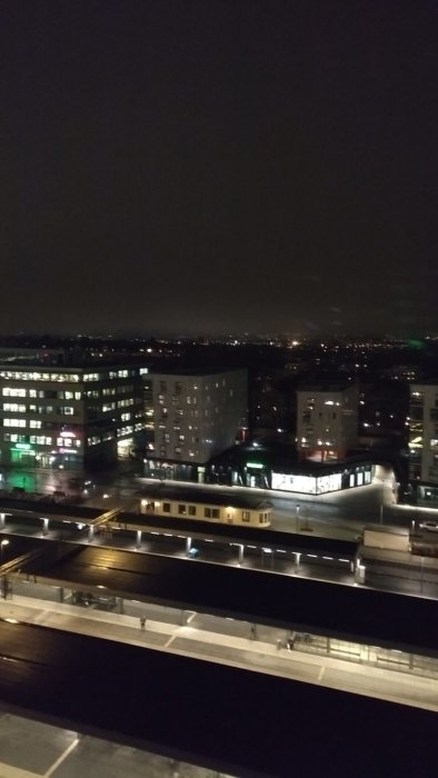 Stadsutsikt på natten med byggnader och tågstation, gatubelysning och diffust ljus på avstånd.