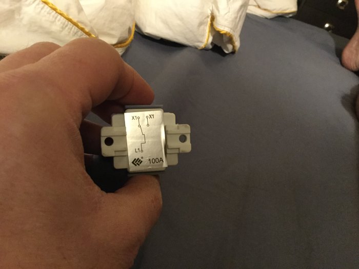 Hand håller en elektrisk trappströmbrytare med etiketter L och X på vit bakgrund.