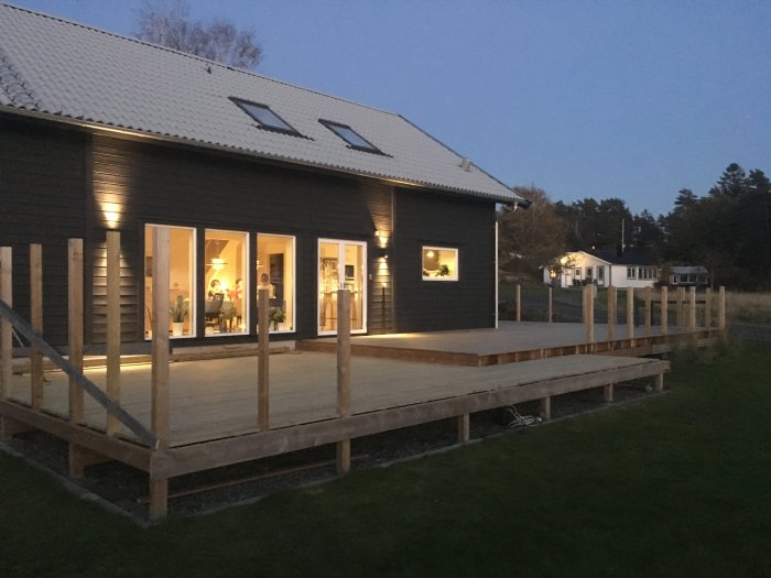 Nybyggd tvånivåers träaltan runt ett mörkt hus i kvällsljus med tända lampor inomhus.