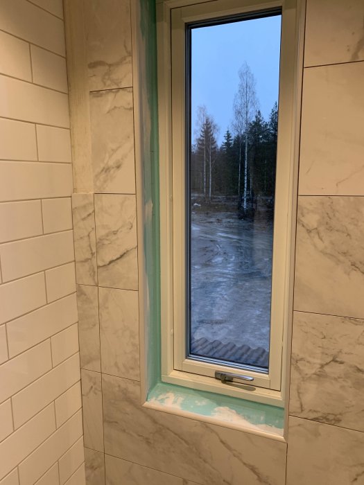 Hörn i ett badrum under renovering med kaklade väggar och synligt spackel vid fönsterkarmen.