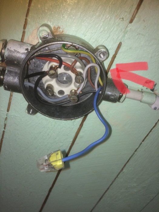 Elanslutning i dosa med synliga skruvanslutningar och en röd pil som visar strömriktning.