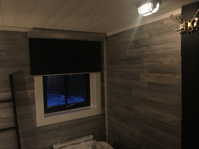 Sovrum med väggar klädda i ljust laminat, mörk rullgardin, svart stege och vit taklampa.