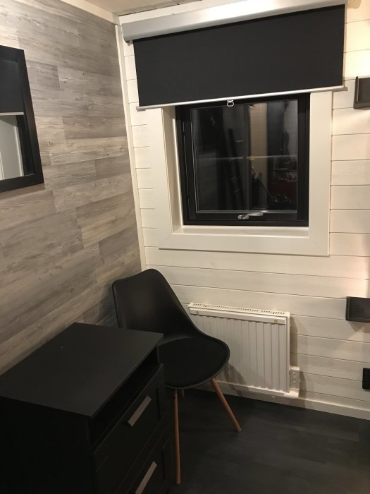 Modern inrett sovrum med golvformat laminat på väggarna, svart skrivbord och stol, mörkt rullgardin för fönstret och vit radiator.