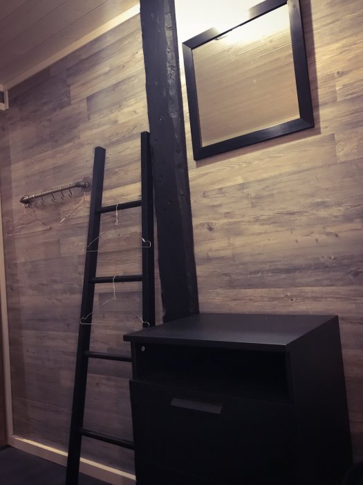 Sovrum med träpanel på väggar, svart stege, svart byrå, en spegel och krokar på väggen.