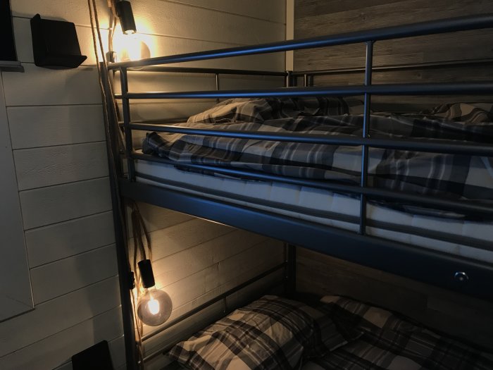 Rustikt sovrum med våningssäng, plädar i rutmönster, lampa och OSB-skjutdörr med läderhandtag.