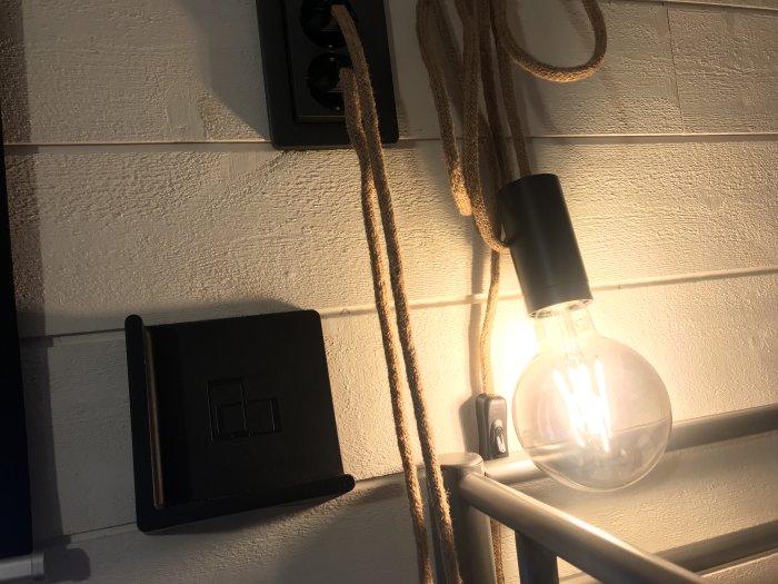 Vägglampa med stor glödlampa och svart armatur, detaljbild av vit strukturtapet och en mobilhållare med laddkabel.