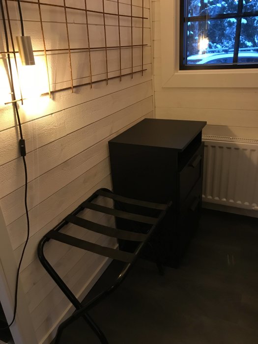 Ett hörn av ett sovrum med svart nattduksbord, vägglampa ovanför och metallbagagehylla, vita panelväggar och mörkt golv.
