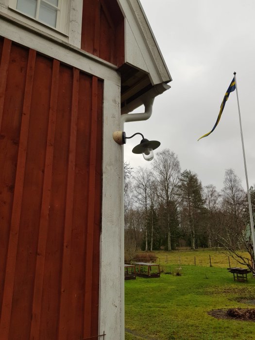 Nyinstallerad utelampa på ett rödmålat husväggshörn med svensk flagga och trädgård i bakgrunden.