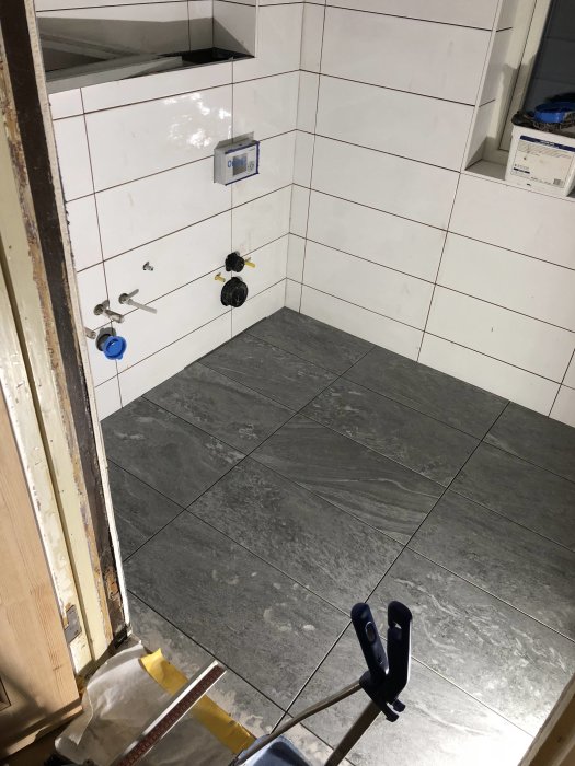 Renovering av badrum med nya vita väggkakel och gråa golvkakel, rörinstallationer synliga.