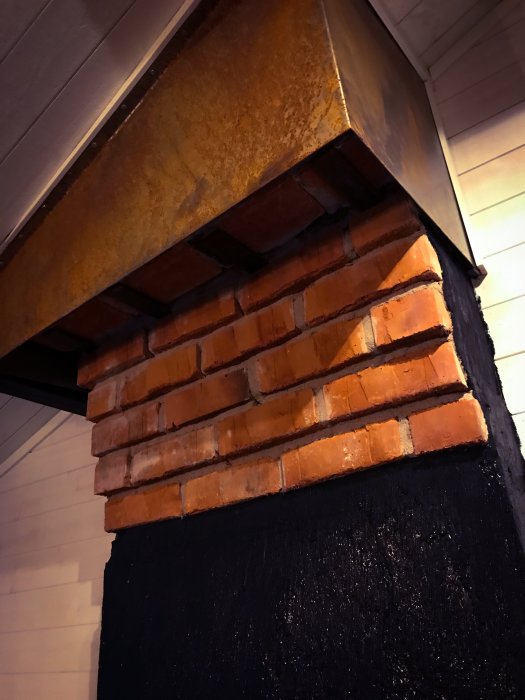 En del av en skorsten i ett renoverat vardagsrum med en rostig plåt ovanpå och tegelstenar målade med svart strukturfärg nedanför.